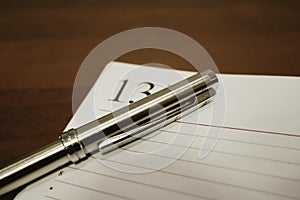 Close-up: a steel pen and an open notebook lie on a brown table with a wooden texture. On the page is the number 13