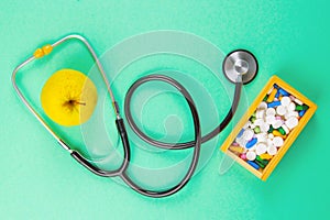 Close-up stethoscope and yellow apple on light green background with pills in wooden box. Top views