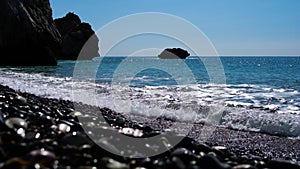 Close-up of stones in sunlight with foamy waves rolling on pebble beach. Blue Mediterranean Sea water crashing on rocks