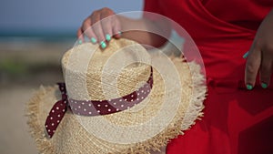 Close-up straw hat with unrecognizable slim Caucasian young woman in red dress sitting on bench outdoors. Happy