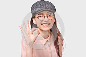 Close up studio image of satisfied young woman wearing trendy gray cap and round transparent glasses, showing Ok gestures.