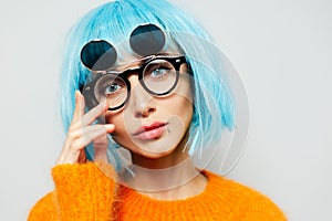 Close-up studio portrait of fashion girl with big blue eyes. Young woman wearing cyan wig, sunglasses and orange sweater on white