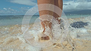 CLOSE UP: Stunning blue waves wash young woman`s sandy legs while on relaxing walk along tropical beach.