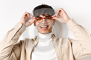 Close-up of stylish hipster guy trying eyewear at optician store, put on glasses and smiling, standing on white