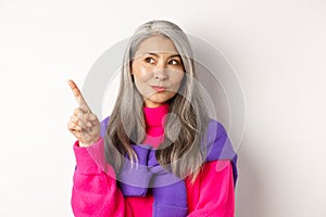 Close-up of stylish korean senior woman with grey hair looking and pointing upper left corner, showing promotion offer