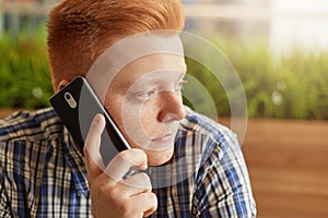 A close-up of stylish redhead man with freckles and dark eyes sitting sideways over greenery holding smartphone on his ear communi