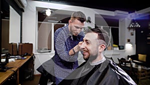 Close up of stylish smiling bearded man sitting covered with black peignoir in the barbeshop. Barber dressed in casual