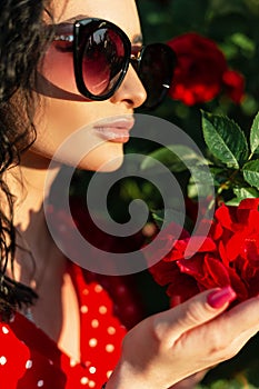 Close-up summer portrait of beautiful woman with fashionable sunglasses in red dress holding a rose in the garden in sunlight