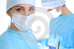 Close up of surgeon woman looking at camera while colleagues performing in background in operation room, isolated