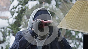 Close-up of suspicious young man in balaclava looking inside house through window. Portrait of Caucasian burglar