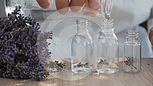 Close-up on the table are several bottles with lavender flowers, a man's hand pours alcohol into the bottles