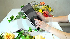 Close-up of tablet on which florist girl`s hands lead fingers among floral arrangements on desk in office in daytime.