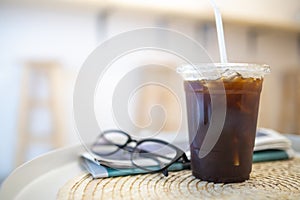 Close up of take away plastic cup of iced black coffee Americano on round table with news paper and reading glasses in the