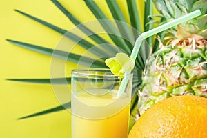 Close up of Tall Glass with Freshly Pressed Pineapple Orange Coconut Juice Straw and Small Flower. Round Palm Tree Leaf on Yellow