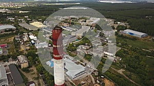 Close-up of tall, red and white chimney-stalk above the industrial zone and factory buildings surrounded by green forest