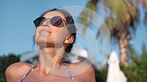 Close-up tanned face of smiling brunette woman in sunglasses enjoying sunbathing in bikini on beach