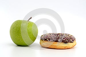 Close-up of tasty chocolate donut and fresh green apple on white background suggesting healthy eating concept