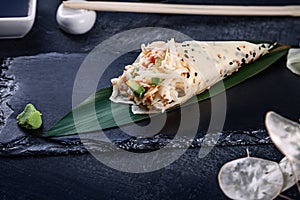 Close-up of tasty hand roll sushi in mamenori with crab and Tobico caviar served on dark stone plate with soy sauce and ginger.