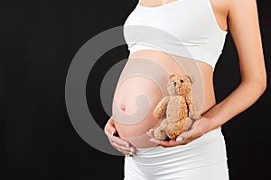 Close up of teddy bear in pregnant woman`s hand at black background. Future mother is wearing white underwear. belly.