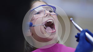 Close-up teenage girl opening mouth as dentist using mouth mirror and dental drill in slow motion. Female adolescent