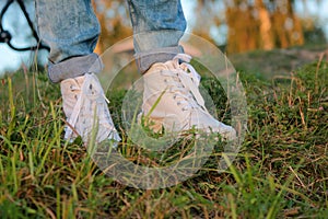 Close up of teenager`s feet in modern and trendy white sneakers and rolled up jeans. Sunny summer day, green grass background.