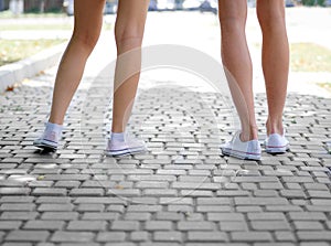 A close-up of teenages` legs in white sneakers standing on a blurred cobbled background. Copy space. Outdoors, yout