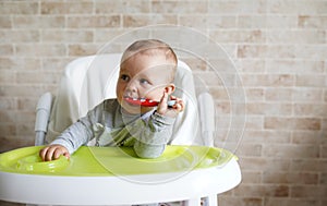 Close-up of teether toddler girl sitting on feeding chair gnawing plastic spoon. Cute infant teething and biting spoon before