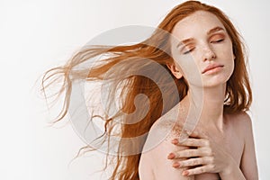 Close-up of tender young woman with long natural red hair flying in air, close eyes and relaxing on white background