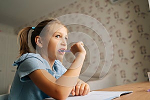 Close-up of thinking primary little child girl doing homework and holding pen against mouth sitting at home table by