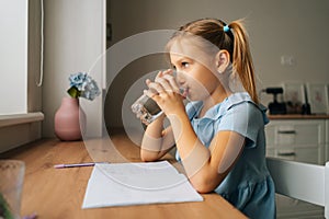 Close-up of thirsty primary little girl writing in exercise book, taking break and drinking fresh cool filtered clear