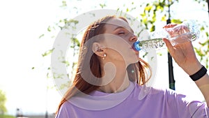 Close-up of thirsty red-haired girl drinking water from bottle outdoors on sunny day.