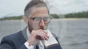 Close-up of thoughtful alcoholic man in eyeglasses drinking from flask sitting on gloomy river bank. Portrait of