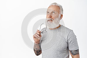 Close up of thoughtful mature bearded man biting temple of glasses, looking up and thinking, making choice and pondering