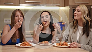 Close-up. Three beautiful girls are sitting at the kitchen table and synchronously take food from the plate with a fork