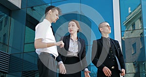 Close up three successful business people stand outdoors near blue glass wall, negotiate, debate, argue. Young confident