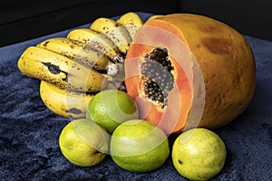 Close up to a fresh sliced orange papaya with seeds inside, a freckled bananas cluster and three green lemons over a blue navy and
