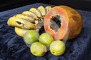 Close up to a fresh sliced orange papaya with seeds inside, a freckled bananas cluster and three green lemons over a blue navy and