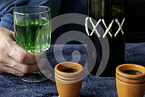 Close up to a man holding a cup of green homemade liquor with two wooden cups and green artesanal bottle photo