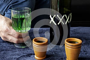 Close up to a man holding a cup of green homemade liquor with two wooden cups and green artesanal bottle