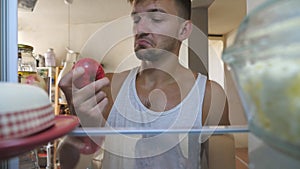 Close up to sleepy man looking for a food at fridge to cooks breakfast. Young lazy guy finding apple on shelf, biting it