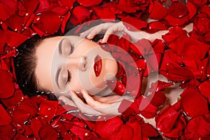 Close-up, top view, beauty portrait, face of a young woman in a bath with water and floating red rose petals and rosebuds.
