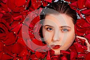 Close-up, top view, beauty portrait, face of a young woman in a bath with water and floating red rose petals and rosebuds.