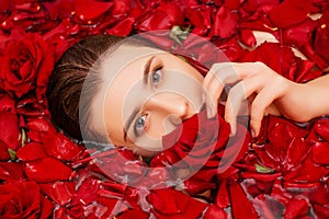 Close-up, top view, beauty portrait, face of a young woman in a bath with water and floating red rose petals and rosebuds.