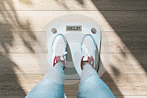 Close up and top view of creative casual dressed feet on scales asking for help. Lose weight and measuring concept. Wooden floor