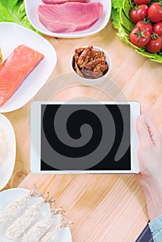 Close-up top view of female hands holding a tablet computer with a blank display surrounded by healthy food on a home