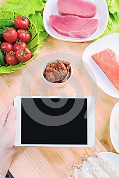 Close-up top view of female hands holding a tablet computer with a blank display surrounded by healthy food on a home