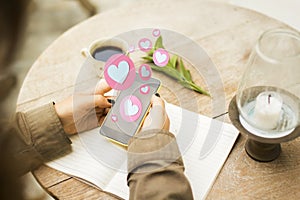 Close up and top view of female hands using smartphone with abstract hearts on blurry background with wooden desktop, candle in