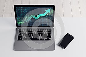 Close up and top view of laptop and mobile phone on office desktop with growing green candlestick arrow. Financial growth, market