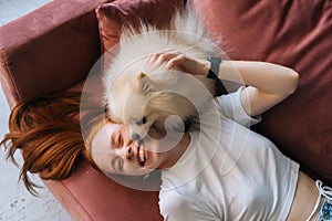 Close-up top view of pretty white small Spitz pet dog lovely licking face of laughing young woman lying on comfortable