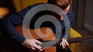 Close-up top view of talented young guitarist man playing guitar sitting on armchair in dark living room at home studio.